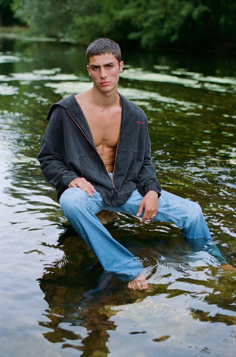 Adult male sitting on a rock in a stream wearing jeans and an open zip up hoodie with a forest and water in the background.