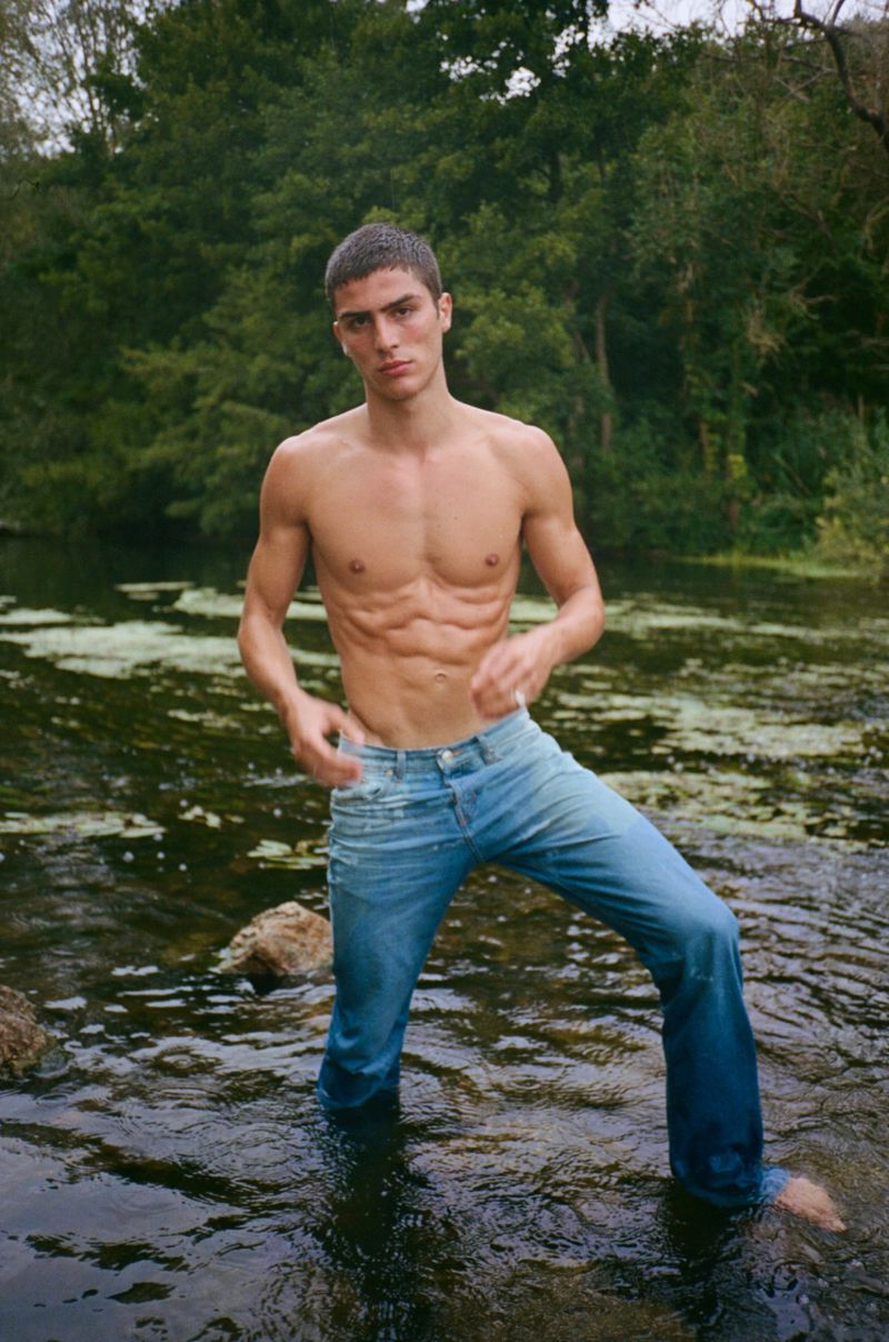 Athletic male model with muscular physique poses in a river with a forest backdrop, wearing jeans, showcasing fitness and outdoor fashion.