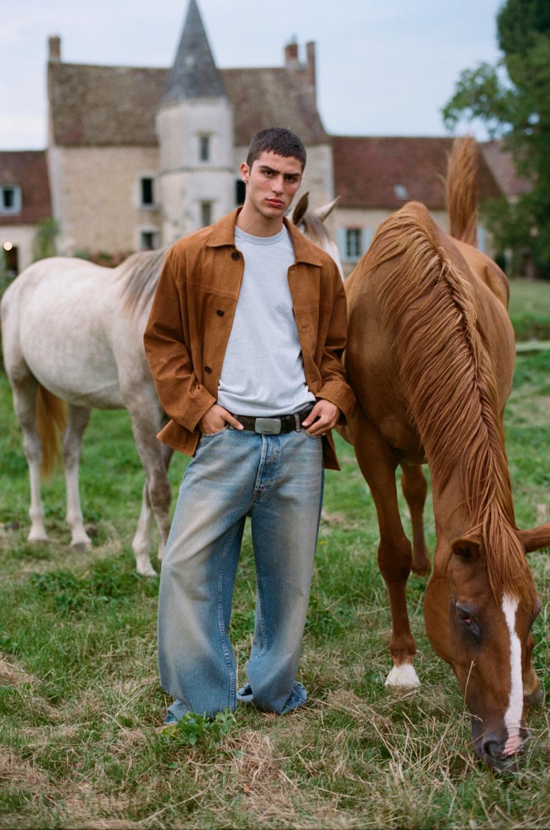A fashionable adult male poses with two horses in a grassy field near a large stone building with a pointed roof.
