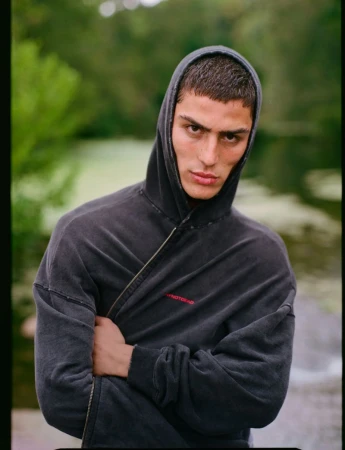 Close-up portrait of a young adult man wearing a hooded sweatshirt with arms crossed, posing in a nature setting with a green background.