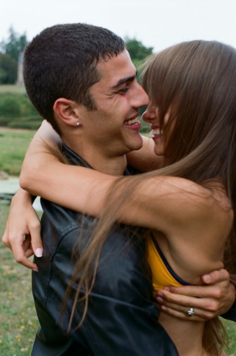 A happy couple is embracing outdoors. The man, with short dark hair, smiles while hugging the woman, who has long brown hair.