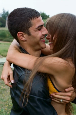 A happy couple is embracing outdoors. The man, with short dark hair, smiles while hugging the woman, who has long brown hair.