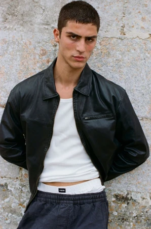 Model Roman Stubrin posing in an open black leather jacket, white tank top, and dark trousers against a textured stone wall.