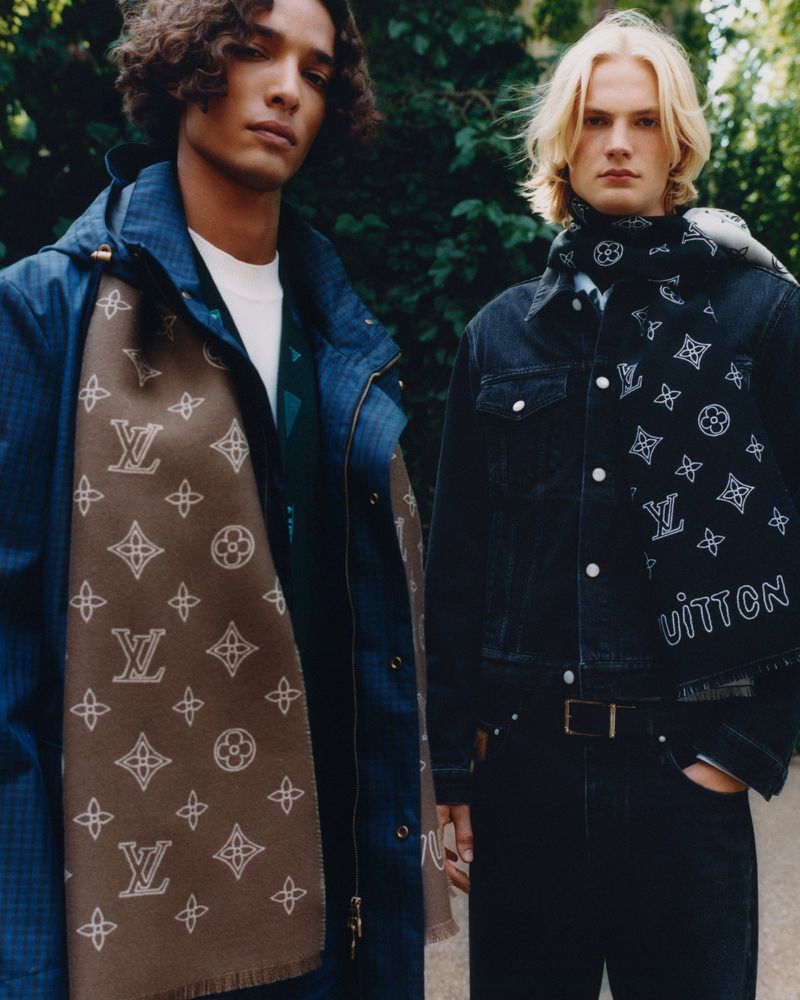 Two adult male models pose outdoors wearing jackets and scarves, each featuring a distinct logo pattern, against a leafy green backdrop.