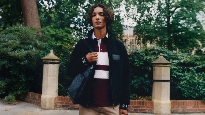 Male fashion model poses outdoors in a polo shirt, jacket, and shoulder bag with a blurred outdoor park and building background.