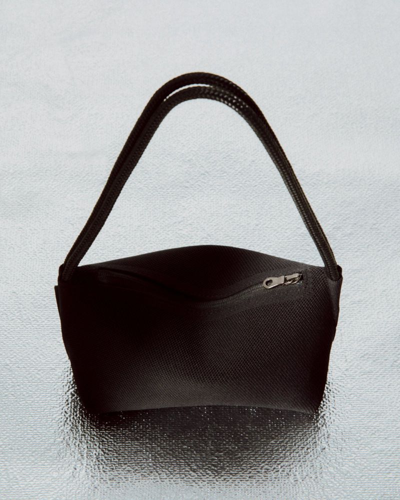 Studio shot of a simple, black fabric handbag with black straps, featuring a zippered pocket, set against a shiny, silver background.
