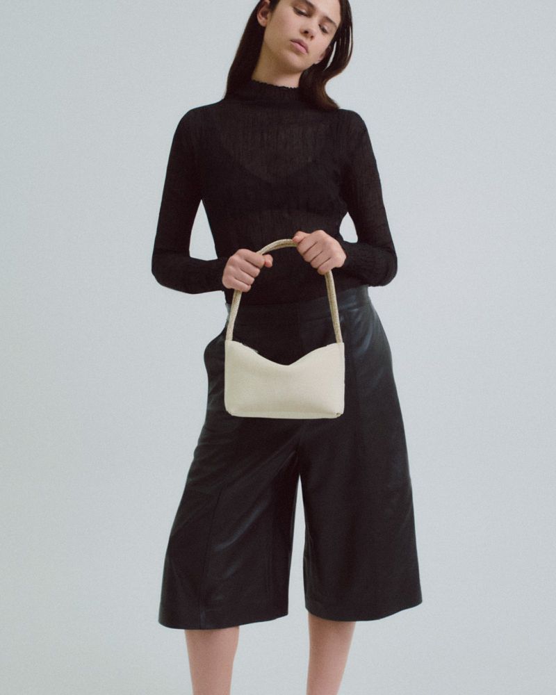 A slender adult female poses in a high-neck black top and black culottes, holding a light-beige handbag against a neutral backdrop.