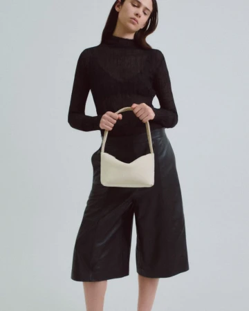 A slender adult female poses in a high-neck black top and black culottes, holding a light-beige handbag against a neutral backdrop.
