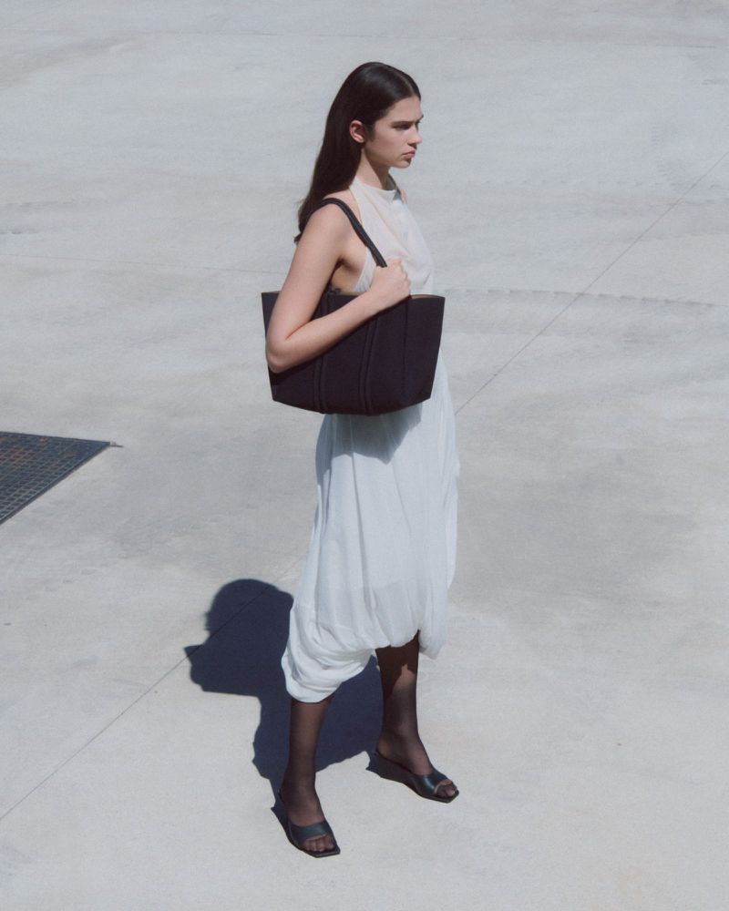 A woman with long brown hair wears a black bag and a white dress while standing on a concrete surface in an outdoor setting.