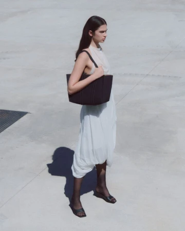 A woman with long brown hair wears a black bag and a white dress while standing on a concrete surface in an outdoor setting.