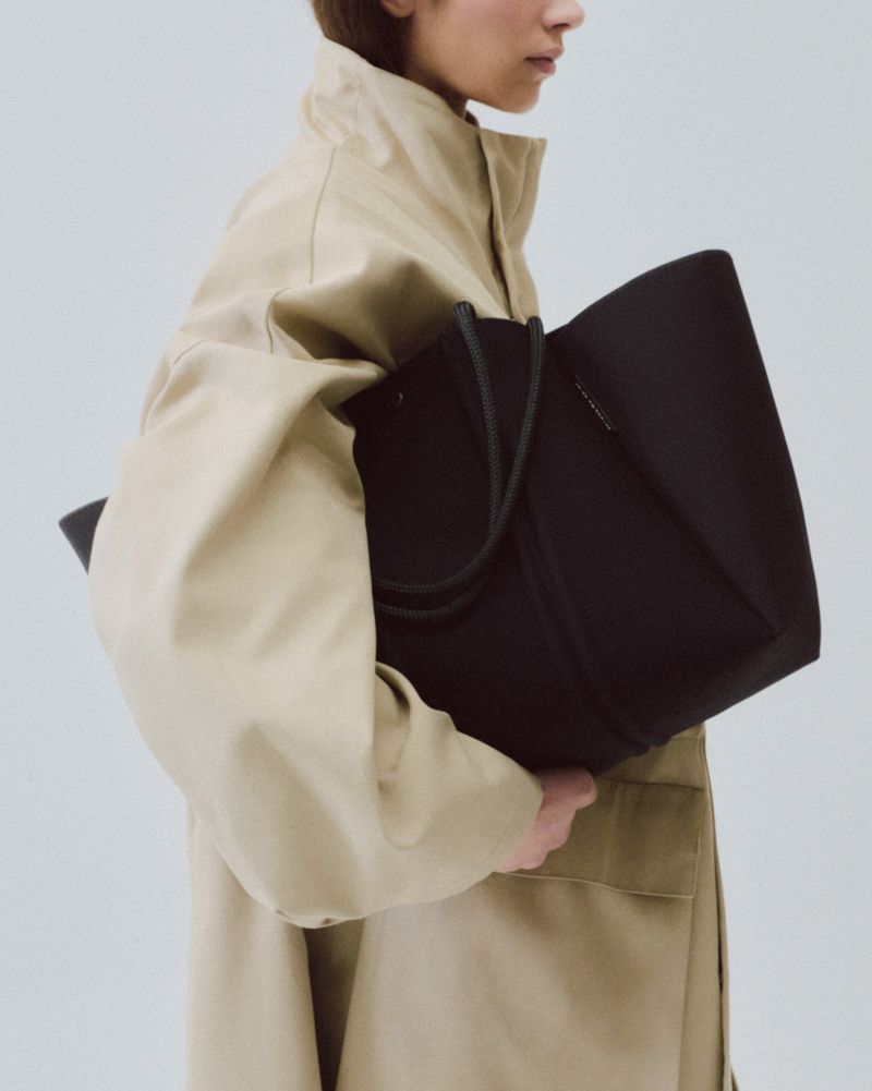 Adult female model carrying a large black tote bag, wearing a tan trench coat in a bright, neutral studio setting.