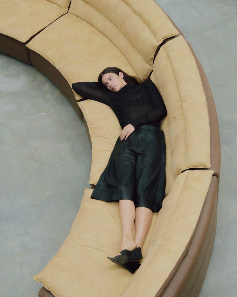 Overhead shot of a woman in black culottes and a sheer top reclining on a tan curved couch, with a smooth grey floor visible.