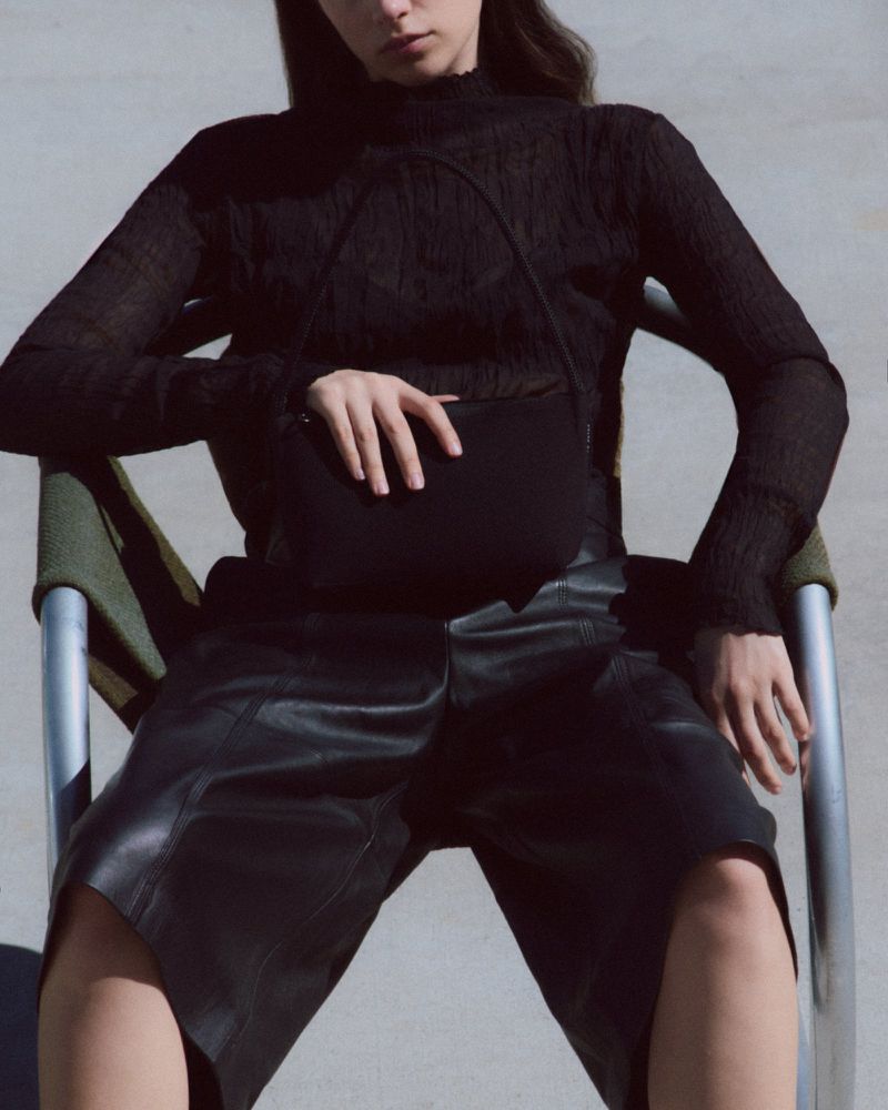 Woman wearing a black textured long-sleeve top and black leather shorts sits in a green chair holding a black purse.