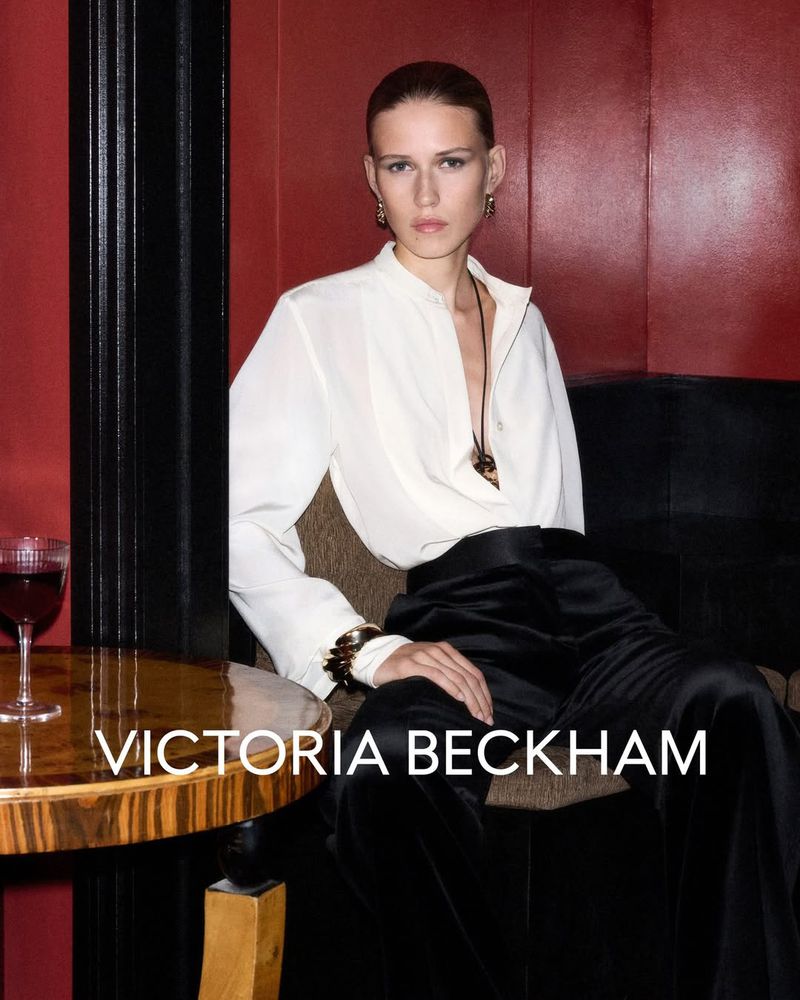Elegant woman in a white top and black pants poses indoors. Glass of red wine and the 'Victoria Beckham' brand are visible.