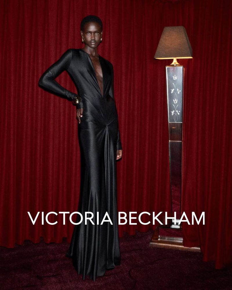 An adult woman poses in a long, black dress next to a standing lamp against a dark red backdrop, showcasing a sophisticated fashion aesthetic.