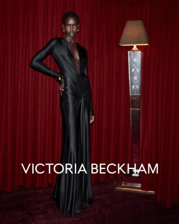 An adult woman poses in a long, black dress next to a standing lamp against a dark red backdrop, showcasing a sophisticated fashion aesthetic.