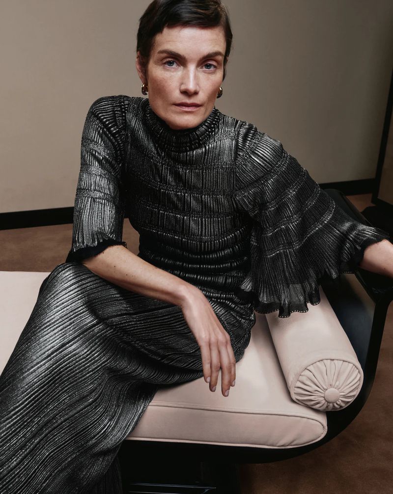 Woman with short hair sits in a metallic pleated high-neck dress on a cream seat, posing for a fashion editorial portrait.