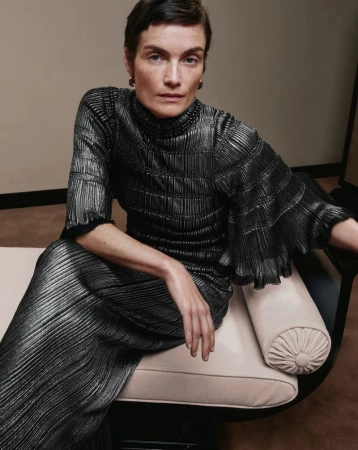 Woman with short hair sits in a metallic pleated high-neck dress on a cream seat, posing for a fashion editorial portrait.