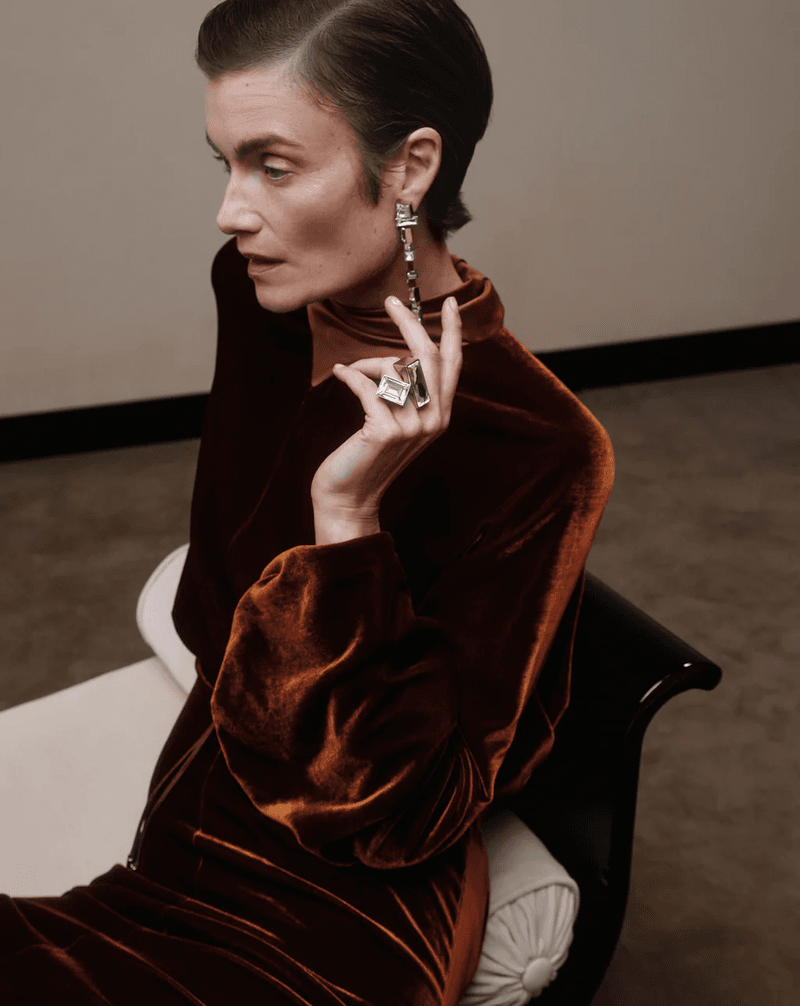 A woman with short hair poses in a brown velvet dress and sculptural jewelry, seated on a white chaise lounge in a neutral studio.