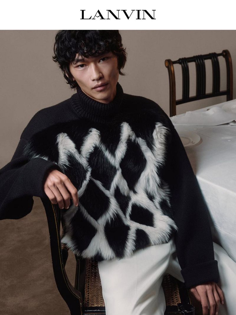 An Asian male model poses indoors wearing a black and white sweater with white pants for Lanvin fashion brand, looking toward the camera.