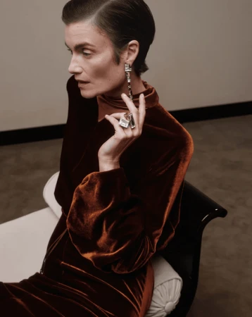 Model in rust-brown velvet top wearing sculptural silver earrings and ring seated against a neutral interior background