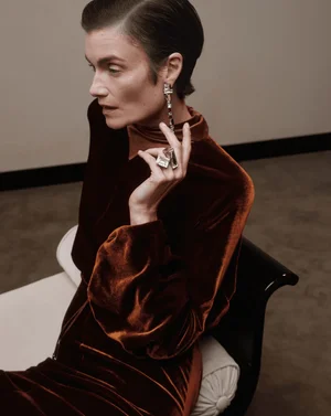 Model in rust-brown velvet top wearing sculptural silver earrings and ring seated against a neutral interior background