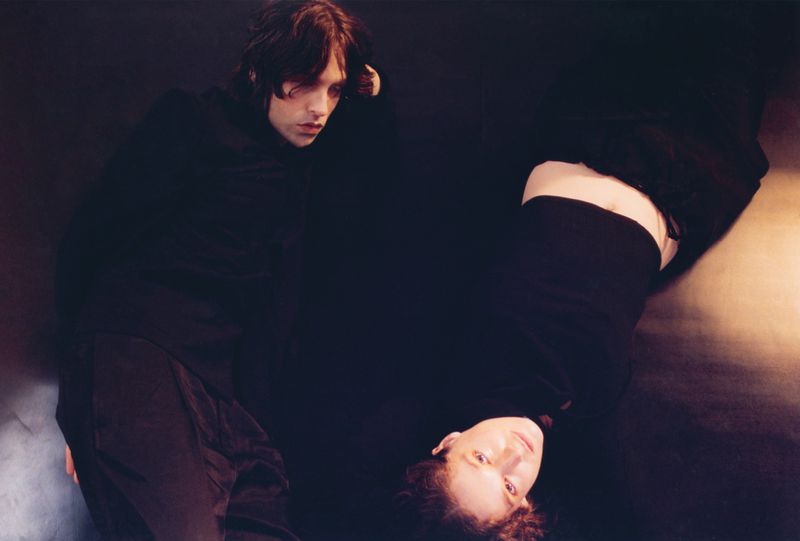 Two people, one light-skinned male and one light-skinned female, pose in a dark studio. He is seated and she is reclining on the ground.