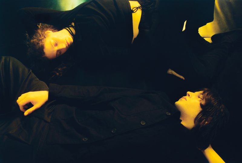 Two people are lying down in the dark in black clothing, close together. Yellow-ish light shines on their faces and hands.