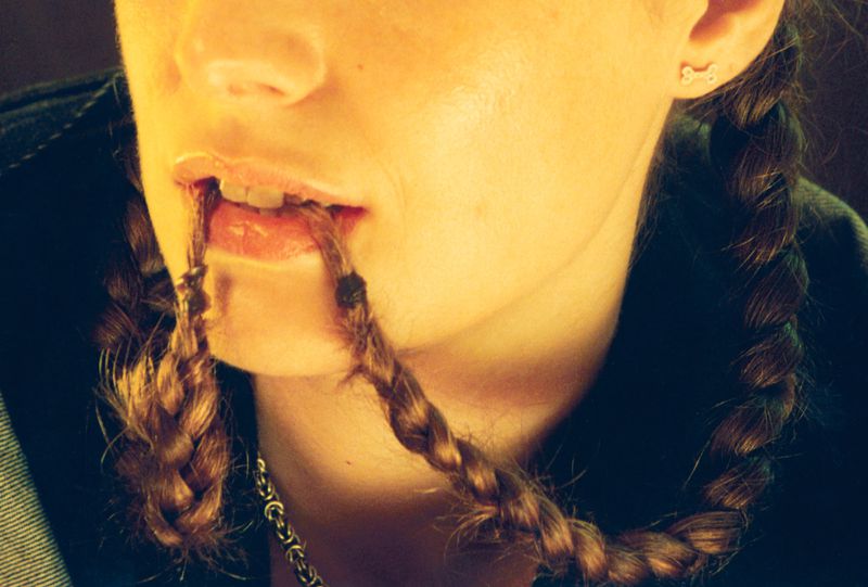 Close-up portrait of a person biting on two braided hair pigtails while wearing a small stud earring and dark jacket.
