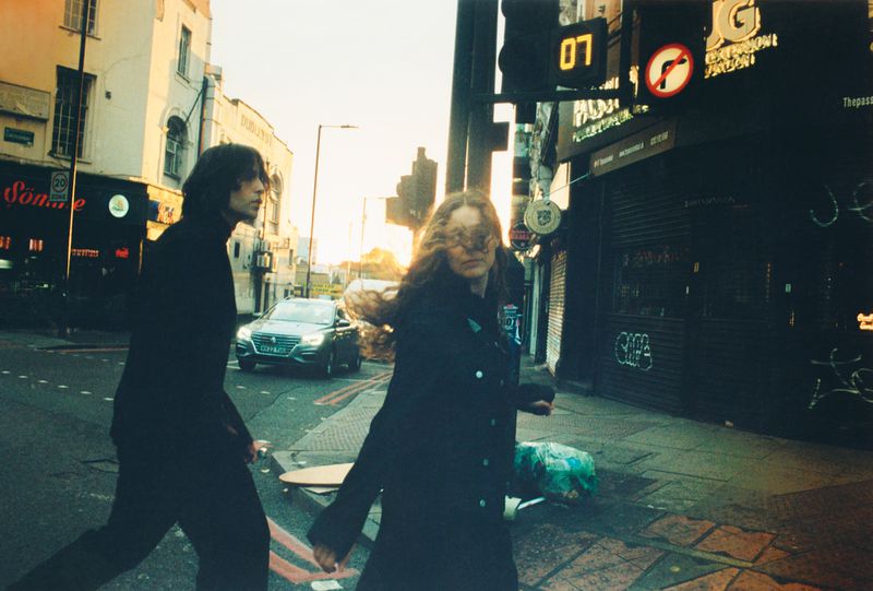 Two adults walk across a city street at dusk near buildings and a stopped car, captured in a wide shot.