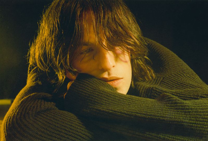 Close-up of an adult with brown hair in a dark green sweater, with hands raised to their face. Low-key lighting and black background.