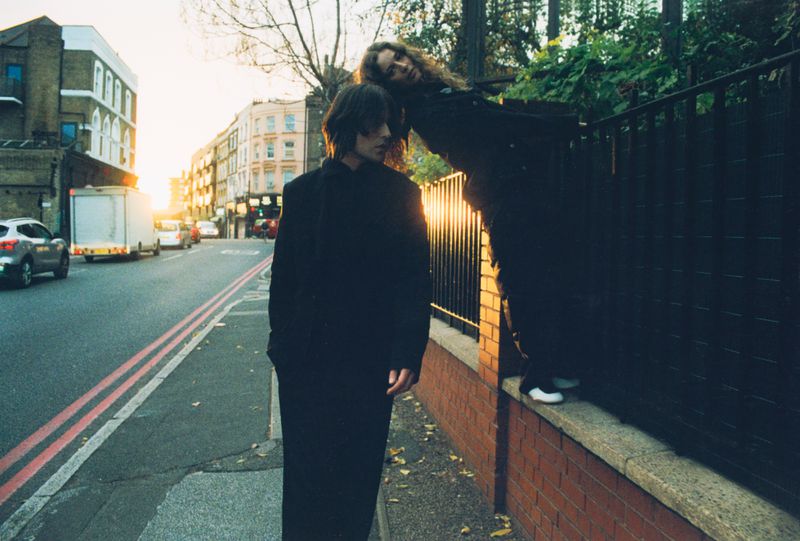 Two people pose outdoors in a city street scene during golden hour, one standing, the other leaning against a brick fence.