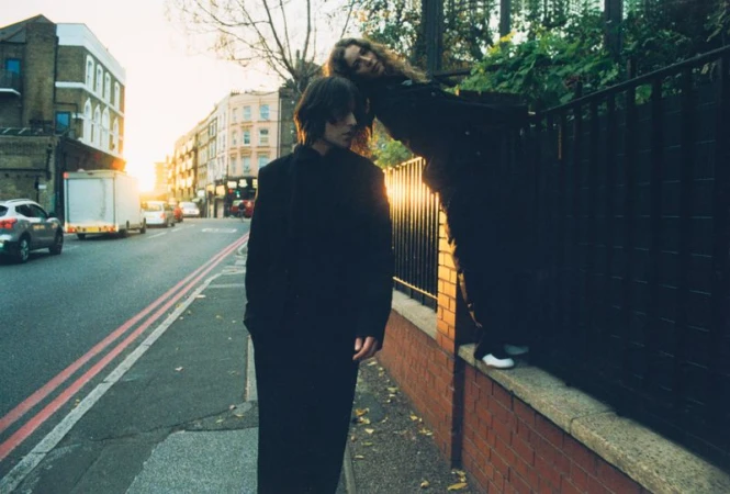 Two people pose outdoors in a city street scene during golden hour, one standing, the other leaning against a brick fence.