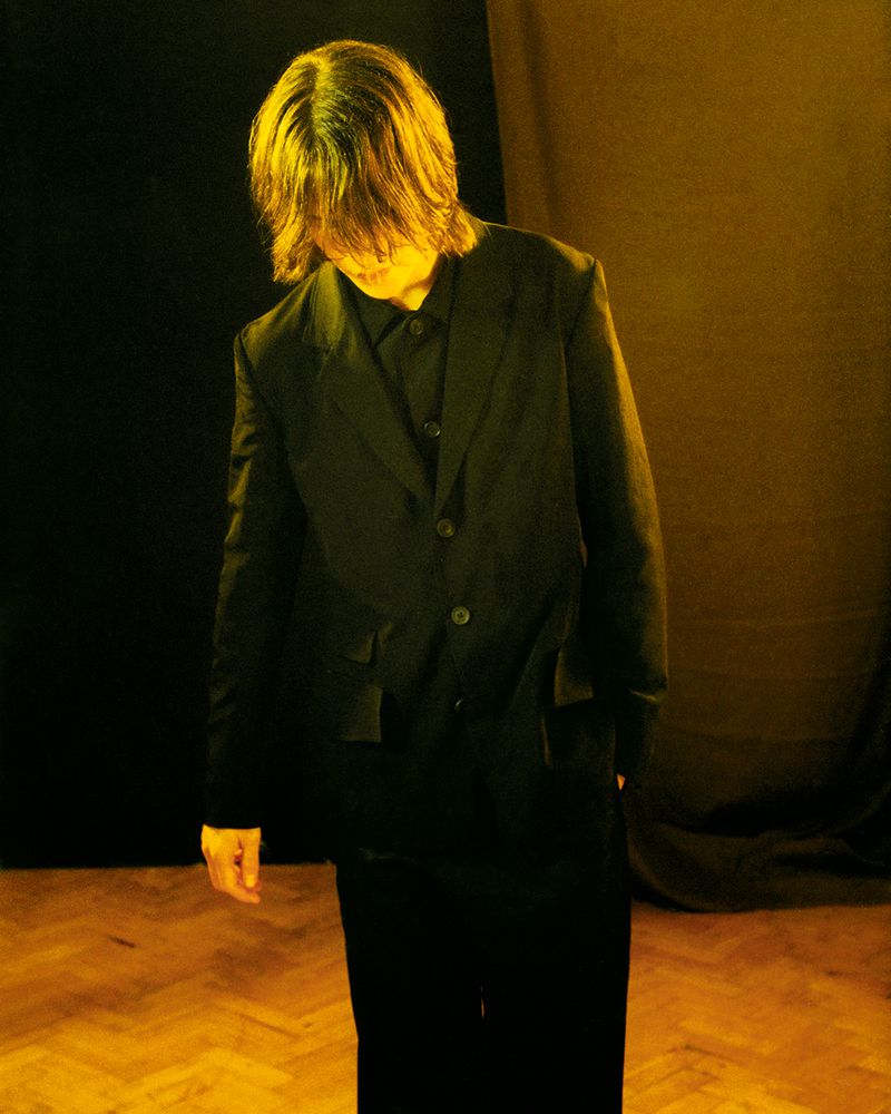 Adult stands in dark indoor setting with wooden floor and yellow lighting, wearing a black suit jacket, shirt, and pants.