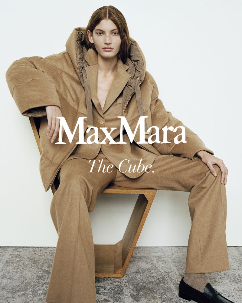 Fashionable adult female model posing while sitting on a wooden chair in a beige hooded jacket and pants for a Max Mara advertisement.