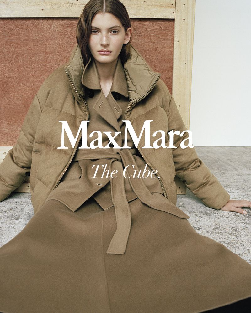 Fashion editorial shot of a woman wearing a tan MaxMara puffer jacket, skirt, and trench coat in a neutral indoor setting with wood paneling.