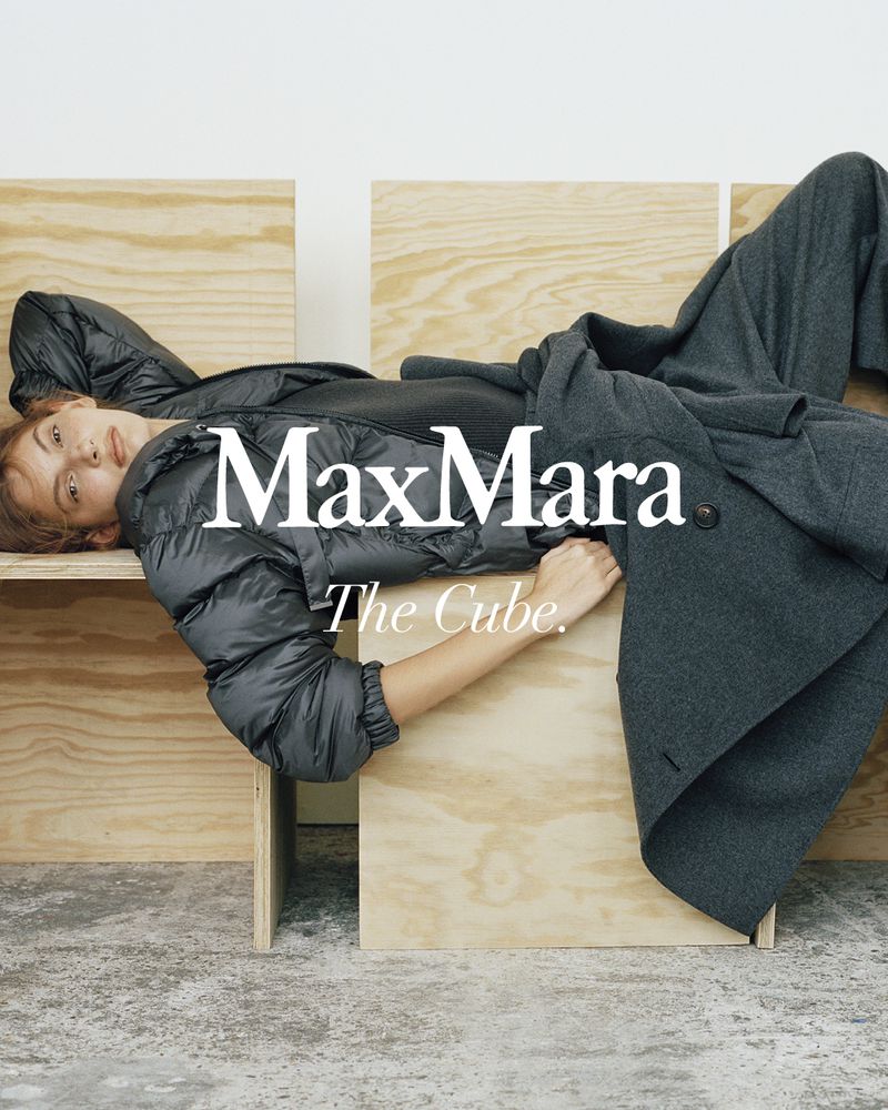 A stylish woman is lying on a wooden cube wearing a black padded jacket and grey wool coat for a Max Mara fashion advertisement.