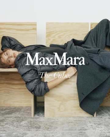 A stylish woman is lying on a wooden cube wearing a black padded jacket and grey wool coat for a Max Mara fashion advertisement.