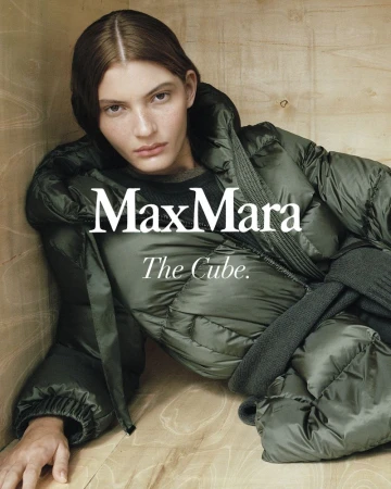 Fashion editorial shot of a brunette woman reclining in a wood cube wearing a green puffer coat, featuring brand logo Max Mara.