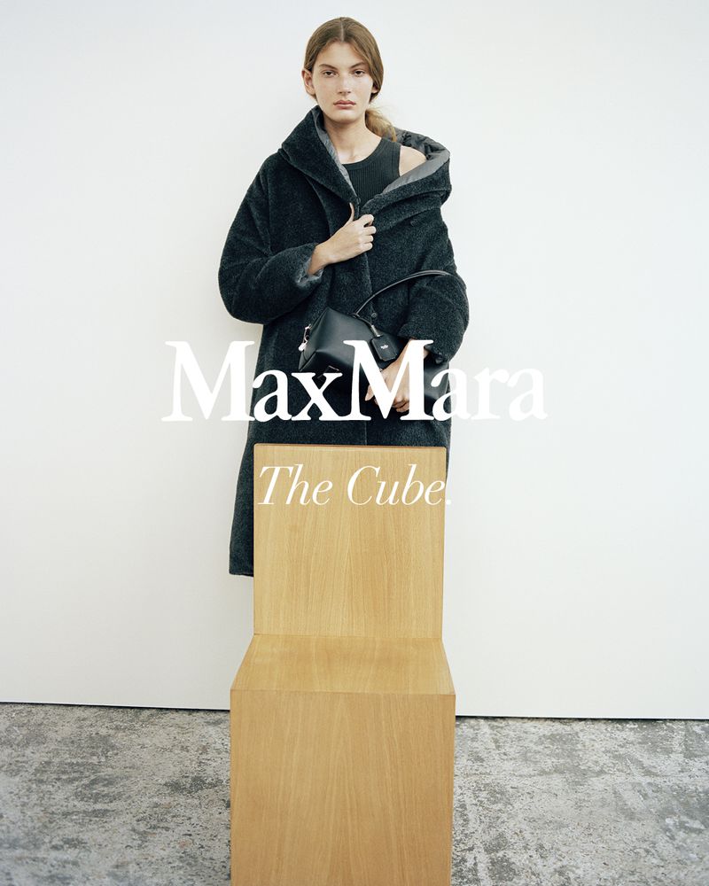 Model wearing a dark textured hooded coat and holding a black handbag, standing behind a wooden chair against a white wall.