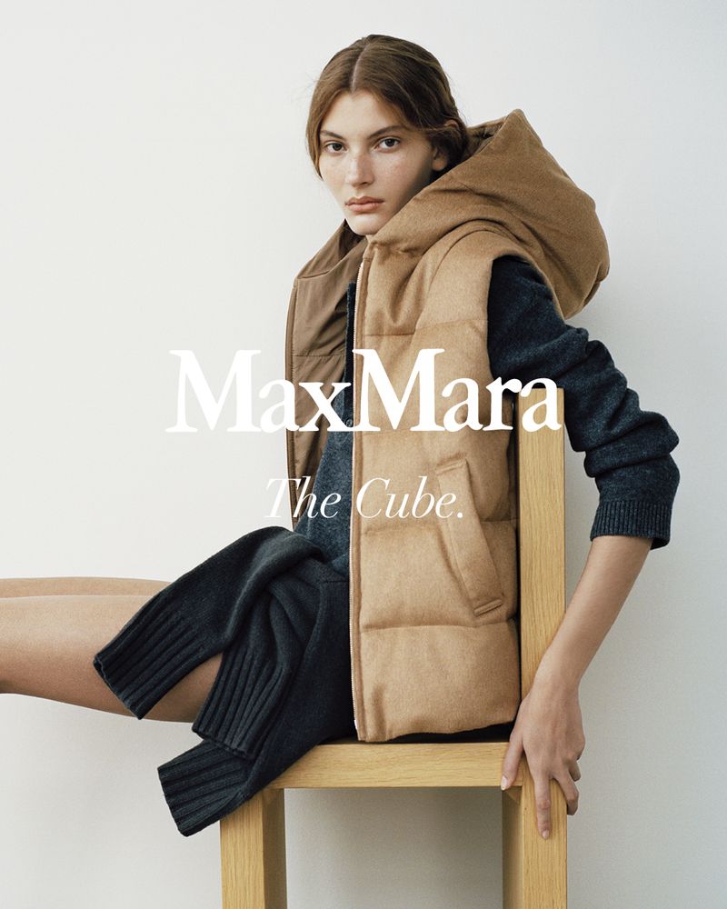 Fashion shot of a woman wearing a Max Mara tan hooded vest and dark grey sweater, sitting on a wooden chair against a white background.