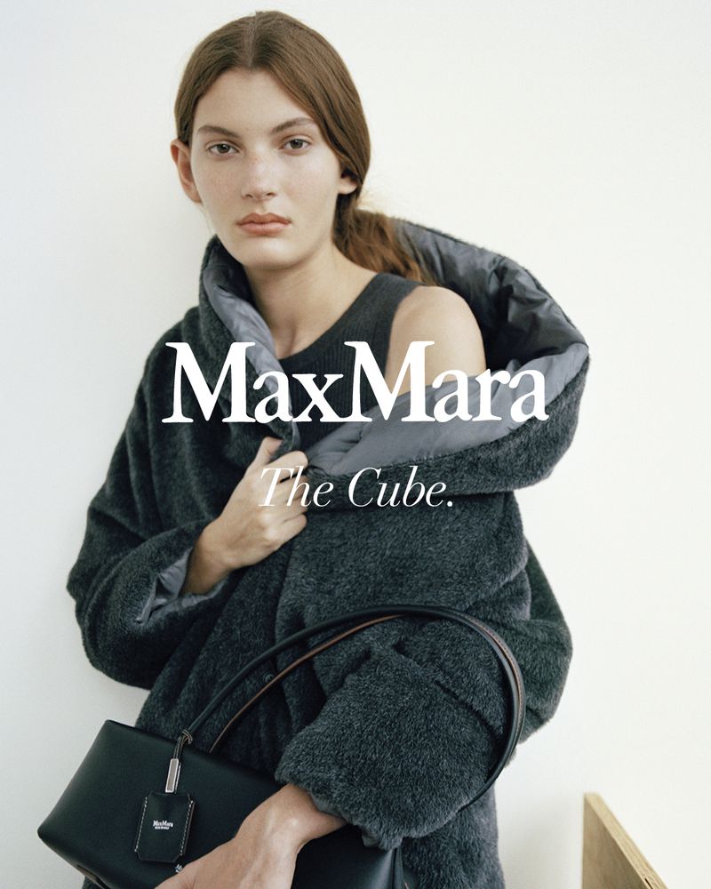 Fashion editorial shot of a fair-skinned woman in a gray, textured coat holding a black, structured handbag indoors against a white wall.