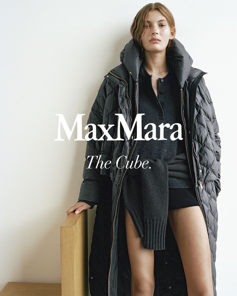 Fashion model in a studio shot, wearing a grey padded long coat and wool sweater, posing against an off-white backdrop with minimal props.