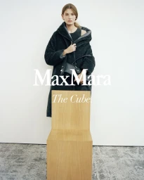 Model wearing a dark textured hooded coat and holding a black handbag, standing behind a wooden chair against a white wall.