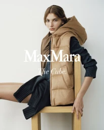 Model seated on a wooden chair wearing a camel hooded puffer vest and dark knitwear against a white studio background.