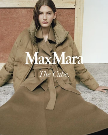 Model wearing a tan belted puffer coat and matching skirt seated against a minimalist architectural background.