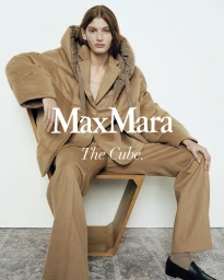 Model wearing a brown camel puffer jacket and matching trousers seated on a wooden chair against a plain white wall