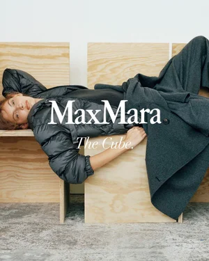 Model reclining horizontally across light wood chairs wearing a black puffer jacket and charcoal wool trousers for Max Mara.