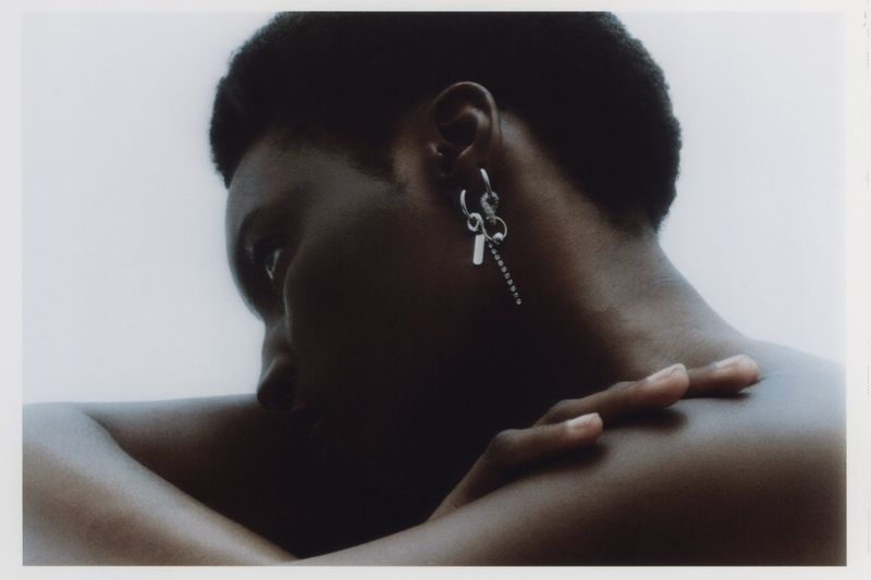 Close-up of Black man with dark hair and a dangle earring posing with arms crossed against a soft white studio backdrop.