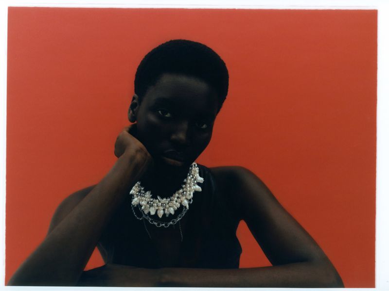 Portrait of a Black female model with dark hair and pearl necklace against a solid red background in a studio setting.